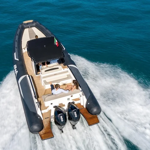 Un grande gommone motorizzato BWA con due fuoribordo Mercury in navigazione sul mare blu. C'è una coppia seduta comodamente in una zona relax sul retro del gommone che si gode la corsa, con un ampio spazio per prendere il sole e rilassarsi.